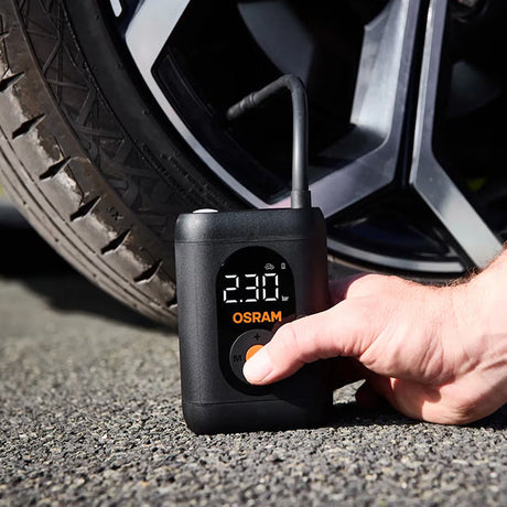 Hånd trykker på tænd/sluk-knap på Osram TYREinflate 1600 ved bildæk, display viser 230 kPa